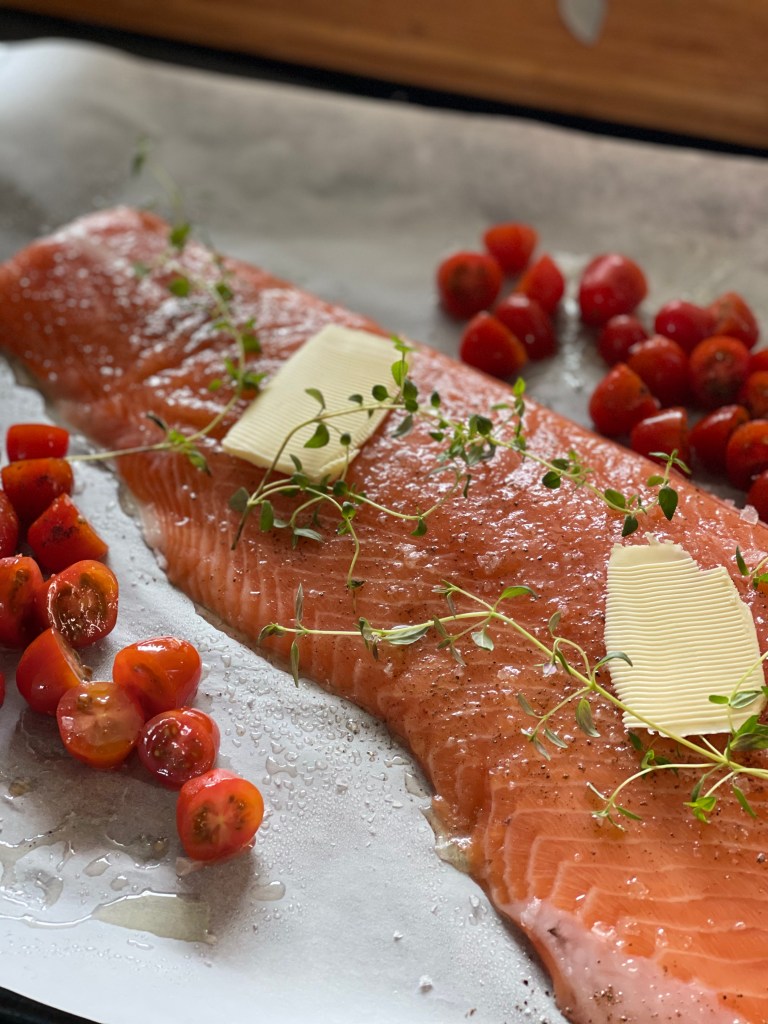 Side of salmon, prior to going into the oven. Lumps of butter, fresh thyme and cherry tomatoes are also visible on the baking tray.