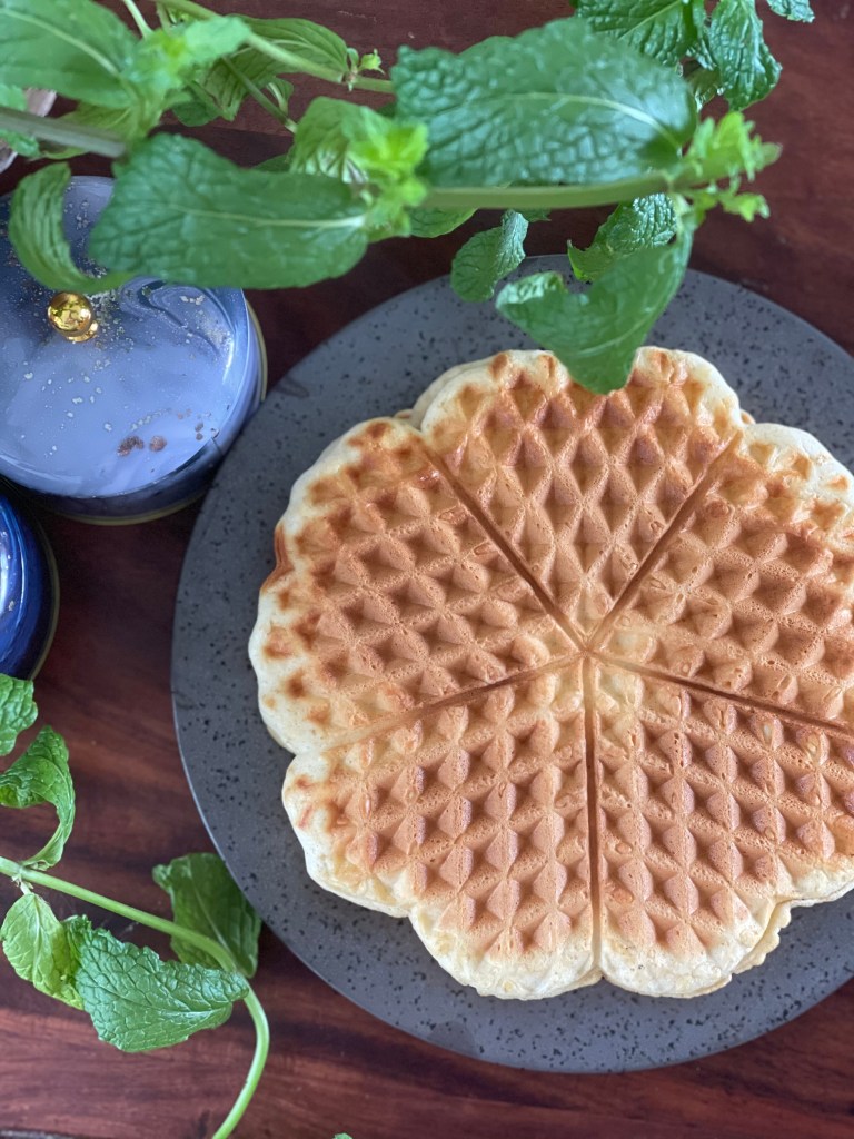 Flatlay of protein waffles, accompanied by a sprig of mint and marbled containers. On a dark wooden desk.