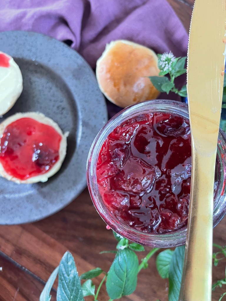 A flatlay display of plum jam in a glass jar. A gilded knife lies diagonally across the top of the jar. Around it, plates of buttered rolls, lush fabrics and green plants create an idyllic image of the jam.