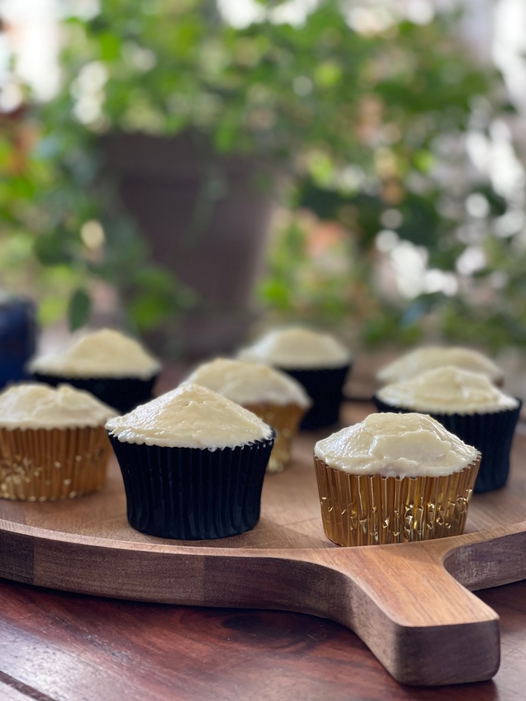 8 cupcakes, alternating in gold and black colors, stand on a wooden board with a lush, green background. It's serene. 